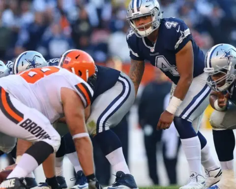 Football players from opposing teams line up for a play; a player in a blue uniform appears ready to receive the snap.