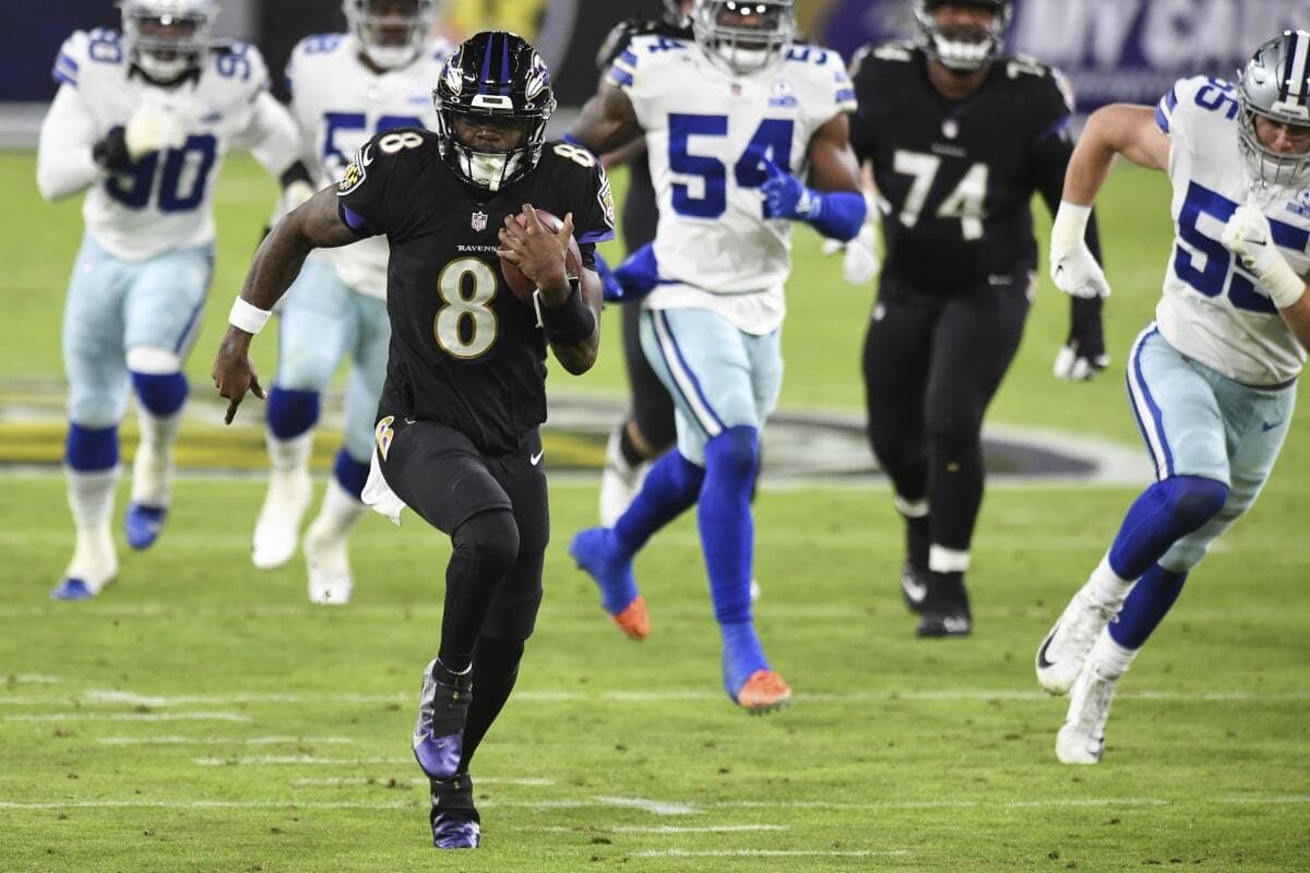 A football player in a black uniform runs with the ball while being pursued by players in white and blue uniforms.