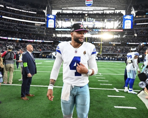 Football player in a white jersey with number 4 walks on the field, smiling, while surrounded by people and press in a stadium.