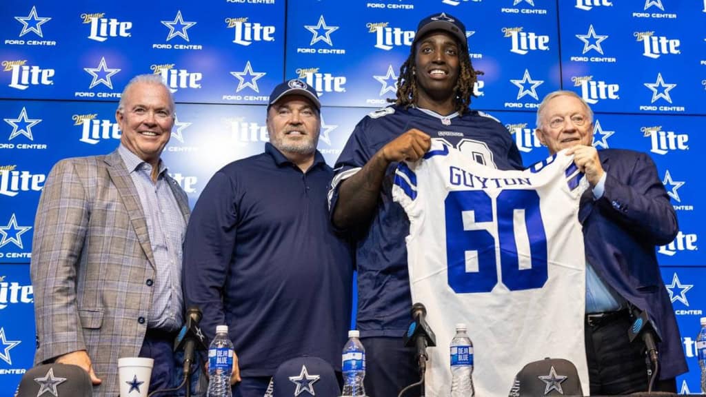 Four men stand together, with one holding up a "Glyton 60" football jersey, at a Dallas Cowboys press event backdrop.