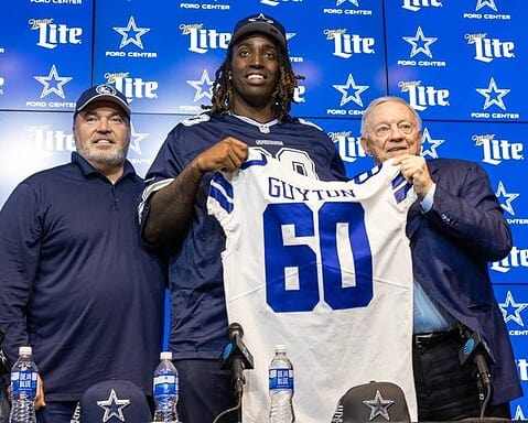 Four men stand together, with one holding up a "Glyton 60" football jersey, at a Dallas Cowboys press event backdrop.