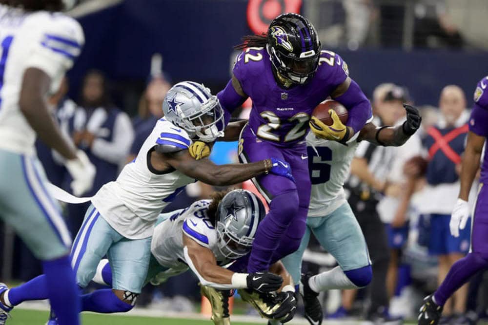 A football player in a purple jersey runs with the ball while two opposing players in white and blue attempt to tackle him.