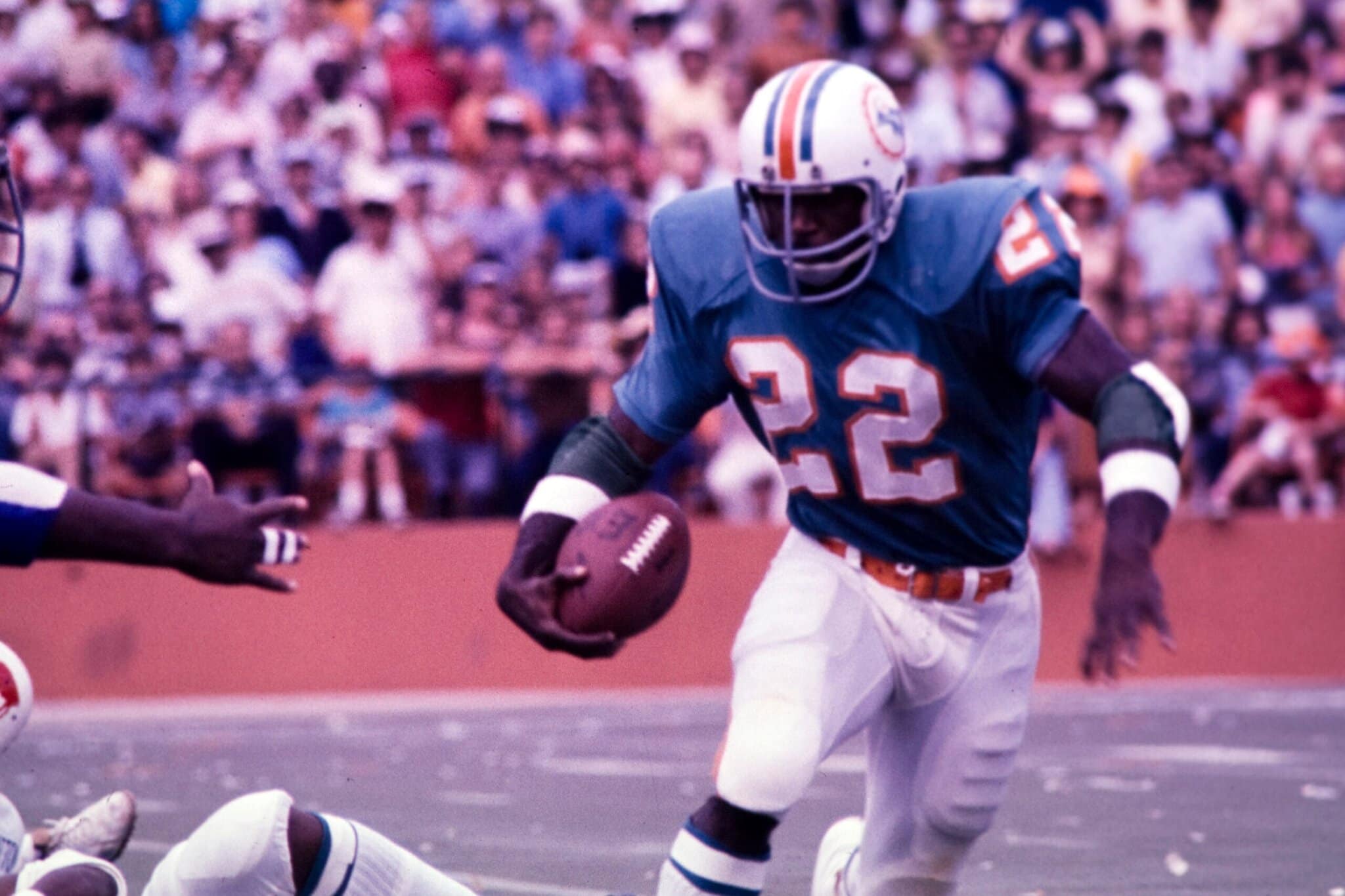 A football player wearing a teal jersey with the number 22 runs with the ball during a game in a stadium.