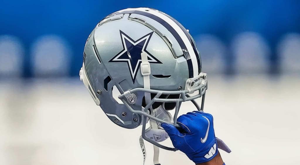 Silver Dallas Cowboys football helmet with star logo, held by a football player wearing blue glove on the field during game day.