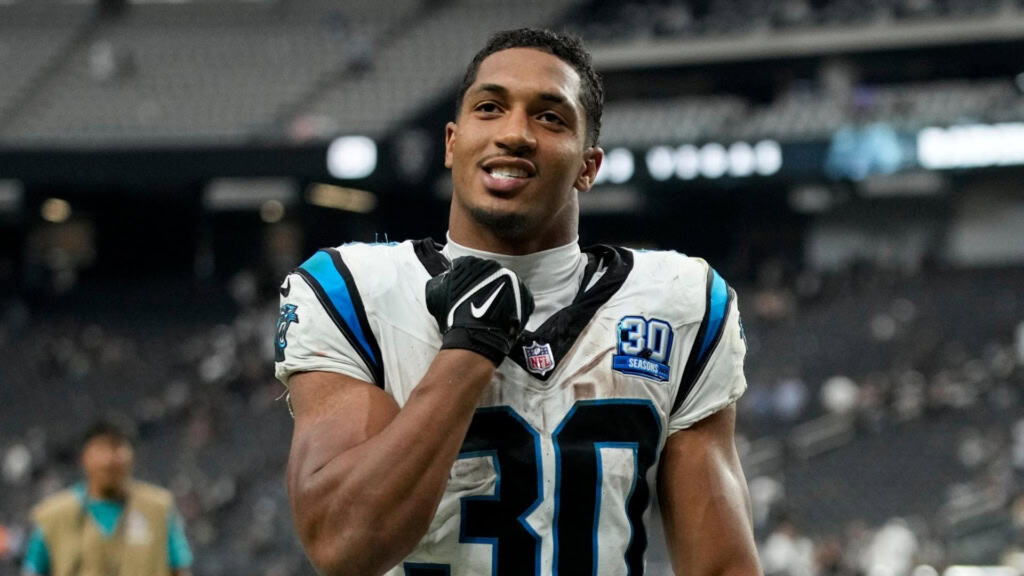 Football player in a Panthers uniform on the field, holding his chin strap and smiling.