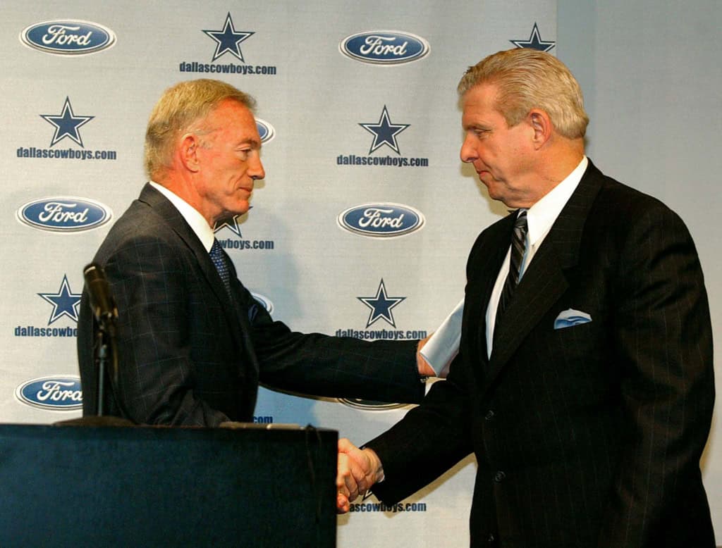 Two men in suits shake hands in front of a Dallas Cowboys backdrop with Ford logos.