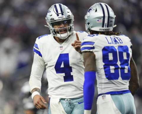 Two football players in white Dallas Cowboys uniforms, adorned with stars, communicate on the field during a game.