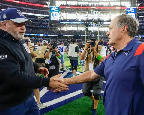 Two football coaches shake hands on the field after a game, surrounded by photographers and staff.