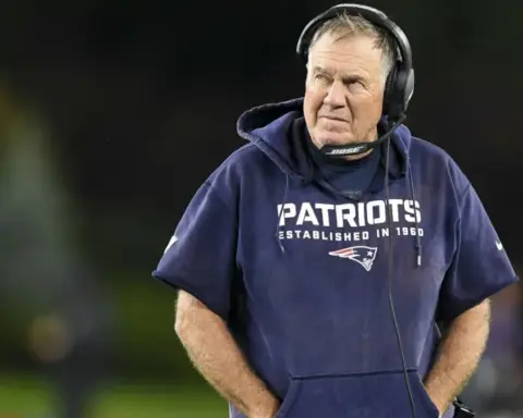 A football coach wearing a Patriots hoodie and headset stands on the field, looking off to the side.
