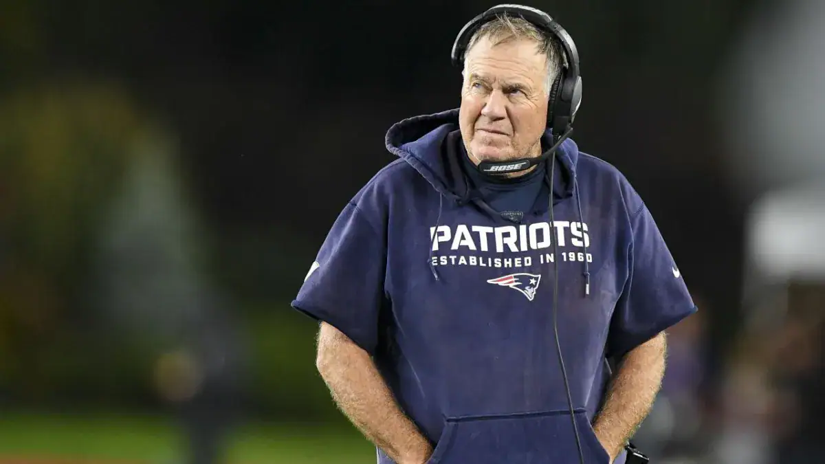A football coach wearing a Patriots hoodie and headset stands on the field, looking off to the side.