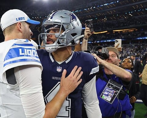 Two football players in uniform shake hands on the field, surrounded by photographers and cameras.