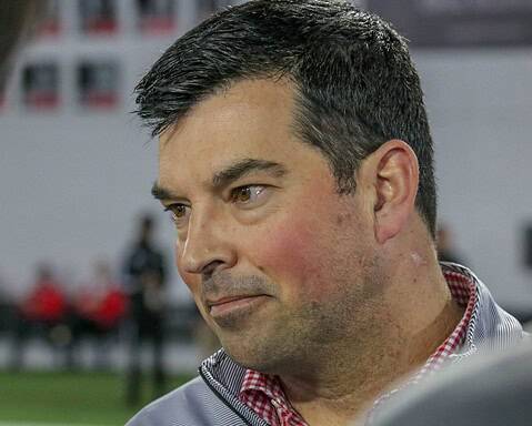 A man in a gray jacket and red checkered shirt looks to the side indoors, exuding the steady focus of a coaching staff member.
