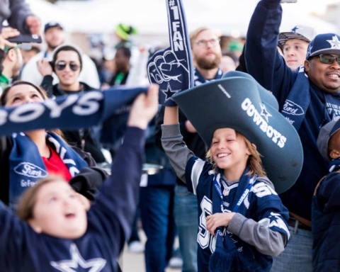 Cowboys fans in team gear cheer enthusiastically at a sports event, waving foam fingers and banners.