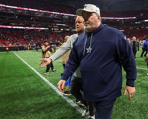 A man in a navy tracksuit and cap walks on a football field, reminiscent of a Dallas Cowboys practice session, with people and stadium seats in the background.