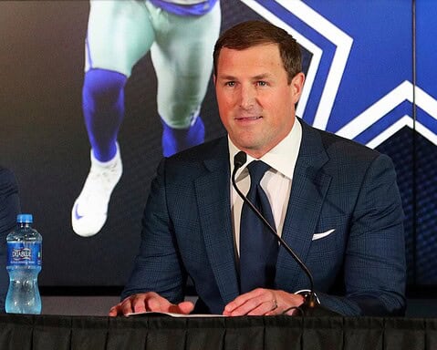 A man in a suit speaks at a press conference, with a sports poster featuring Cowboys legend Jerry Jones in the background and a water bottle on the table.