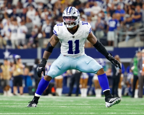 A Dallas Cowboys football player in uniform braces for a play on the field, eyeing the Falcons with determination, as fans glance nervously at the injury report.