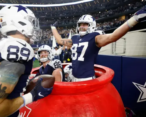During a Thanksgiving game in the 2024 season, a Cowboys football player celebrates exuberantly in a large red bucket on the sidelines, with two teammates laughing nearby.