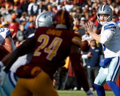 The football player in the white jersey, looking slightly upset, prepares to throw a pass as a determined Commanders player in a red jersey rapidly approaches.