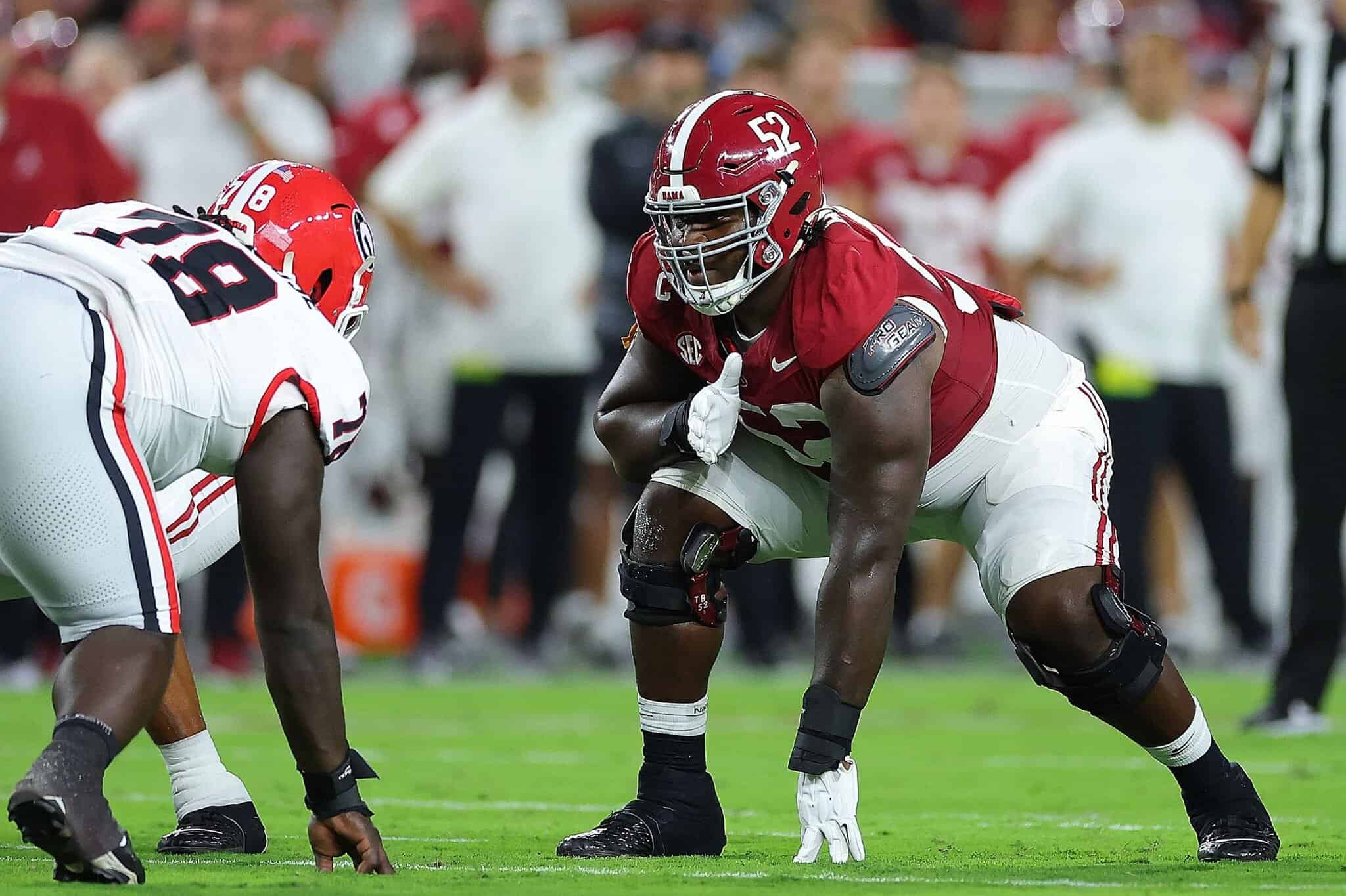 Aggressive football player in red uniform crouching in a defensive stance on the field with teammate in white readiness for play, college football game at night.