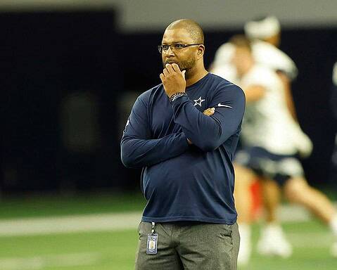 Dallas Cowboys coach standing on the sideline during a game, wearing a navy blue team shirt and glasses, appearing thoughtful and focused.