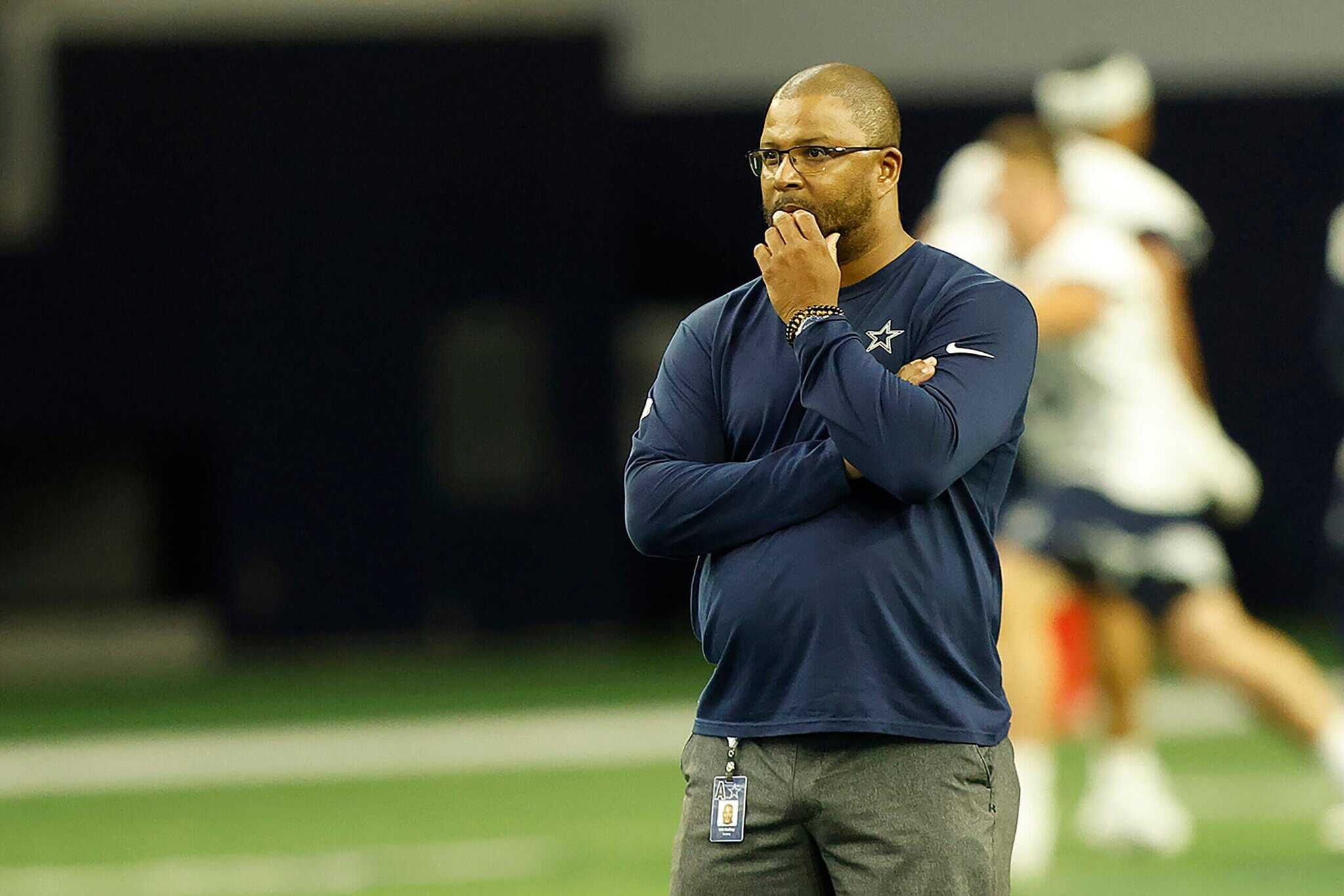 Dallas Cowboys coach standing on the sideline during a game, wearing a navy blue team shirt and glasses, appearing thoughtful and focused.