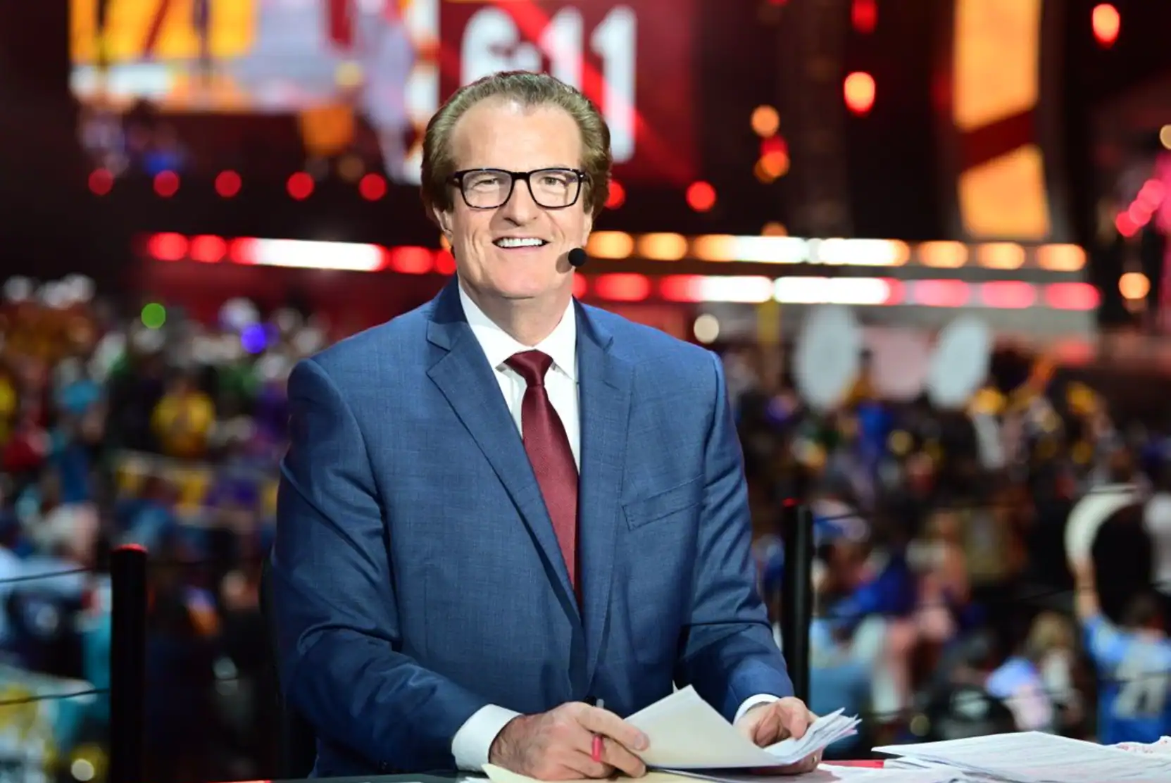 Hosting multiple colorful screens and lights in the background at an indoor event, a smiling male sports commentator wearing a blue suit, white shirt, maroon tie, and glasses, with a microphone headset, providing live coverage of a Dallas Cowboys game.