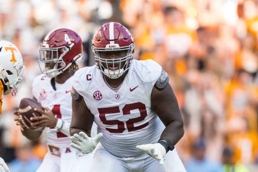 Alabama football player in white jersey with maroon '52' running on the field during game against Tennessee Volunteers, focused on gaining yards, stadium crowd blurred in background.