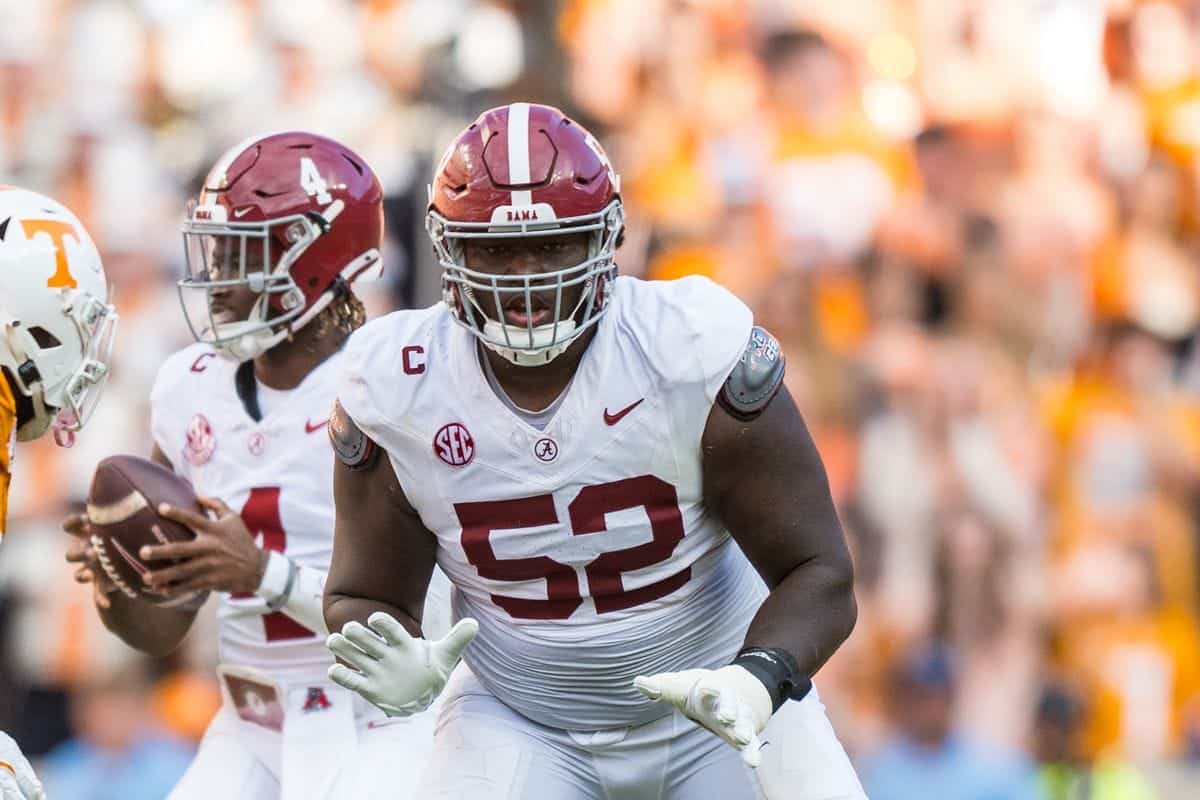 Alabama football player in white jersey with maroon '52' running on the field during game against Tennessee Volunteers, focused on gaining yards, stadium crowd blurred in background.