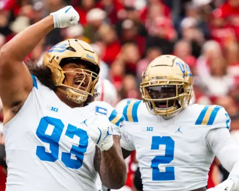 Celebrating U-Mass football players in white uniforms with blue and gold helmets, high-fiving and cheering on the field during a college football game.