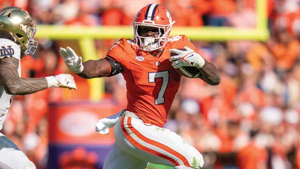 Running back in orange jersey with number 7 carrying football during college football game, defensive player approaching.