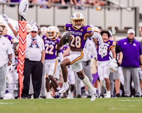 High school football player wearing purple and gold uniform with the number 28 running with the ball during a game, with coaches and teammates on the sideline.