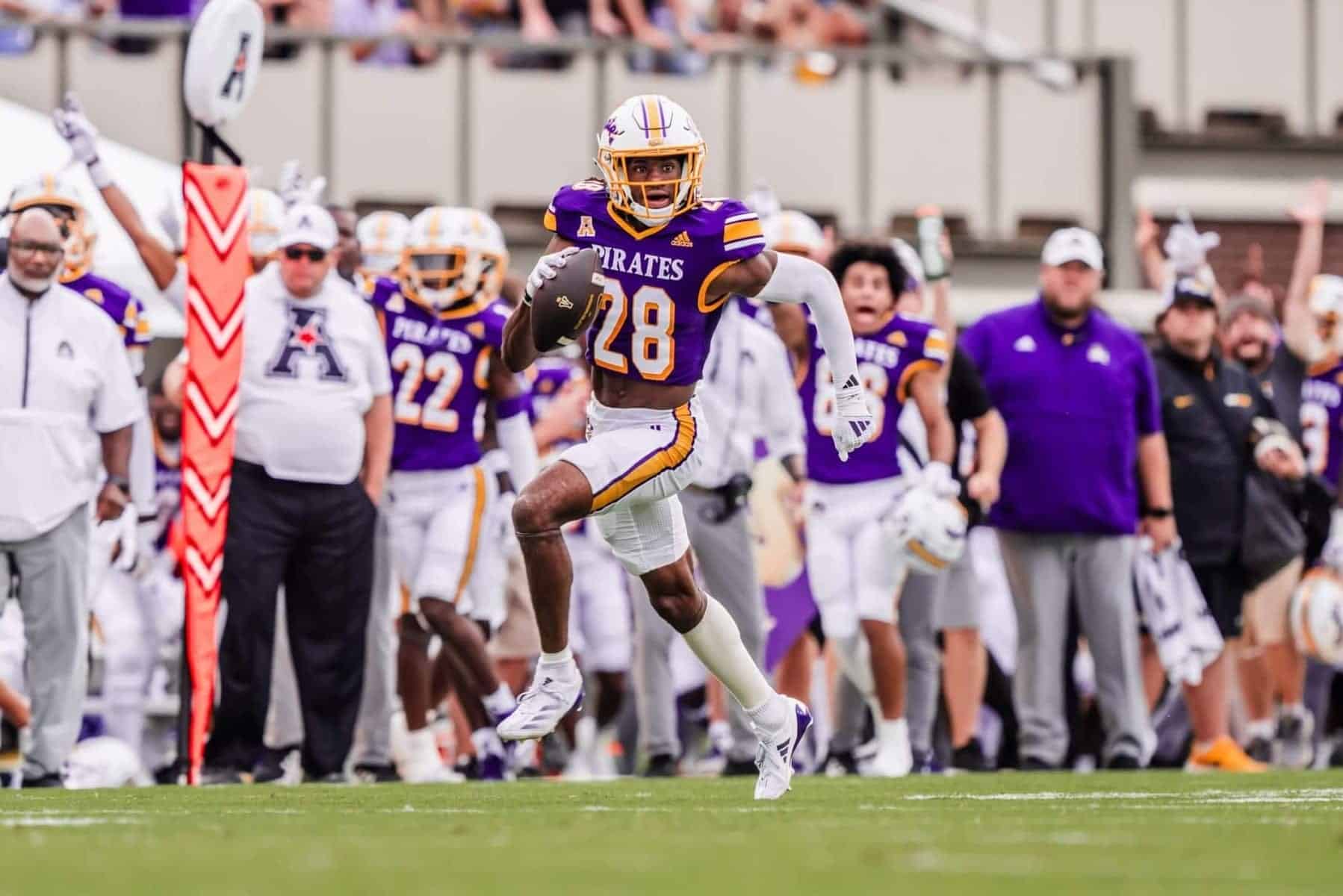 High school football player wearing purple and gold uniform with the number 28 running with the ball during a game, with coaches and teammates on the sideline.