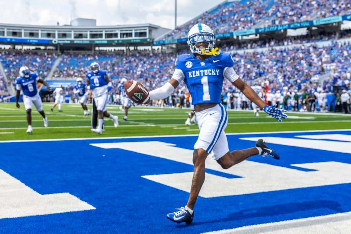 Vibrant image of Kentucky Wildcats football player celebrating touchdown on field during game at Kroger Field, with passionate team and fans in the background, emphasizing college football excitement and sports achievement.