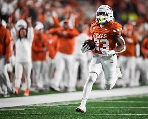 Running back player in Texas Longhorns football uniform carrying football during game at the stadium with cheering fans in background.