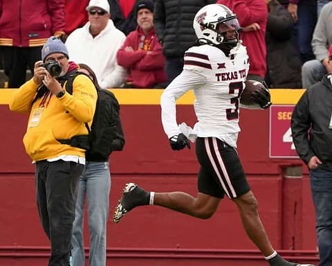 A Texas Tech football player running with the ball during a game at the stadium, surrounded by fans and photographers, showcasing college football action and athleticism.