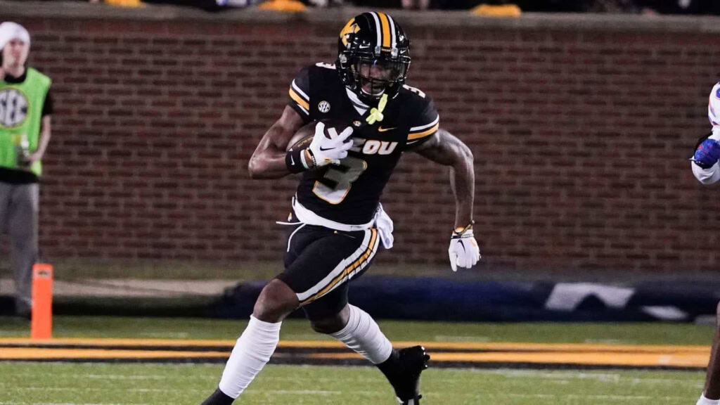 Oklahoma State University football player running with the ball during a game, wearing black and gold uniform, with a brick wall background, focusing on college football action.