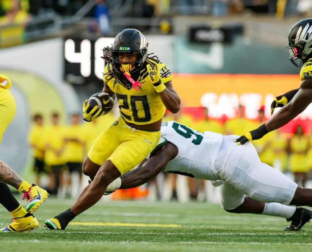 High school football player in yellow uniform running with ball, being tackled by opponent in white jersey during game; action shot with stadium background.