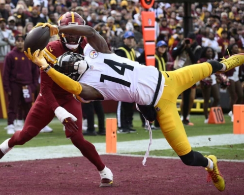 Athletic football player making a catch in an NFL game, wearing the Washington Football Team uniform with a Pittsburgh Steelers defender attempting to block, on the field during a game, with crowd in background.