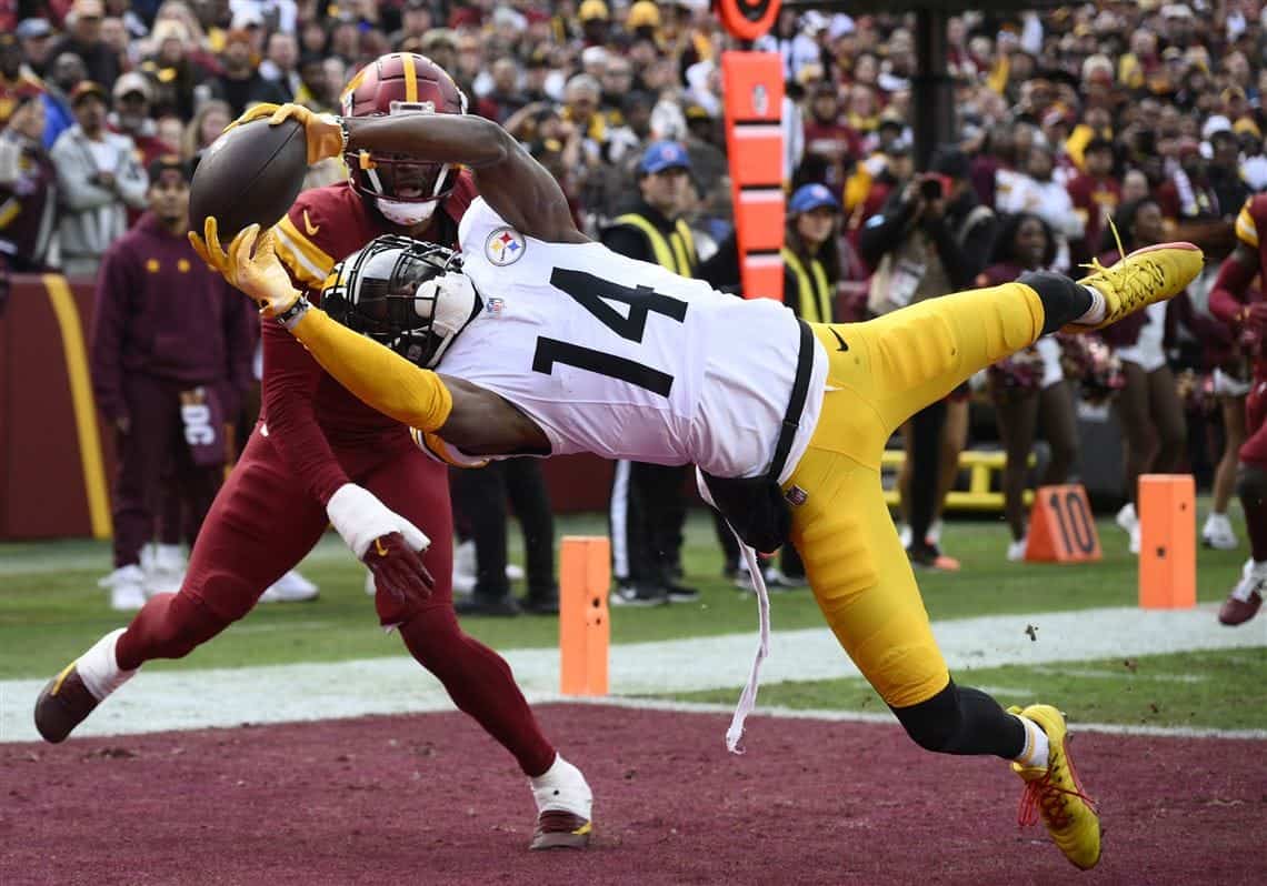 Athletic football player making a catch in an NFL game, wearing the Washington Football Team uniform with a Pittsburgh Steelers defender attempting to block, on the field during a game, with crowd in background.