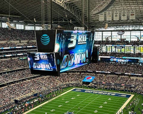 Vibrant indoor football stadium during a Dallas Cowboys game, featuring a large digital scoreboard showing "3rd DOWN," crowded stands, and team branding, highlighting excitement and sports entertainment.