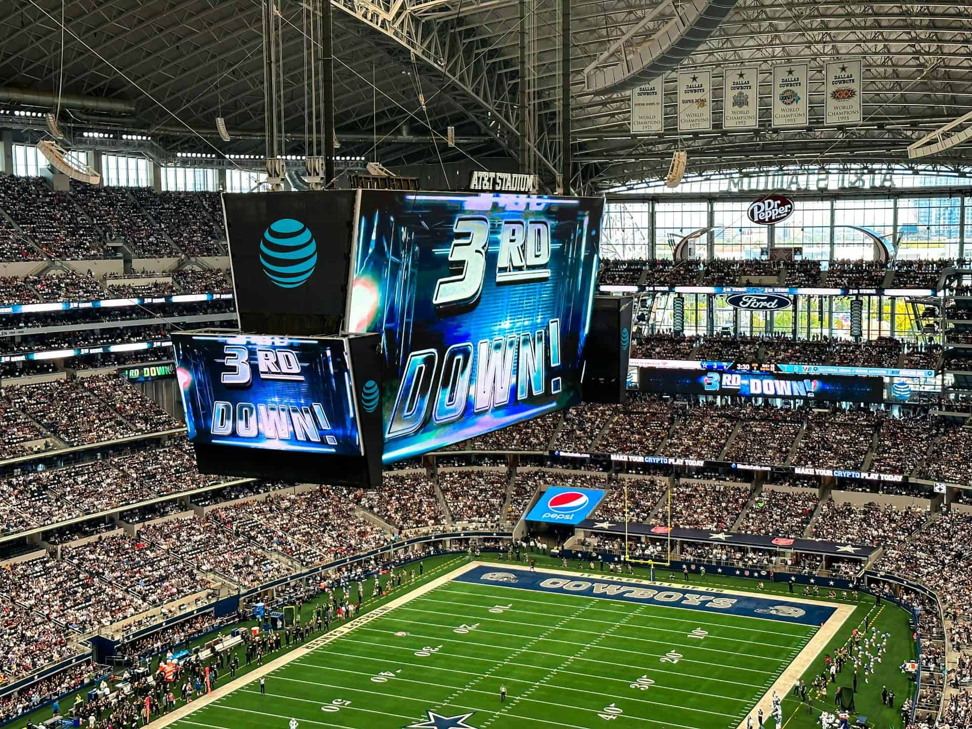 Vibrant indoor football stadium during a Dallas Cowboys game, featuring a large digital scoreboard showing "3rd DOWN," crowded stands, and team branding, highlighting excitement and sports entertainment.