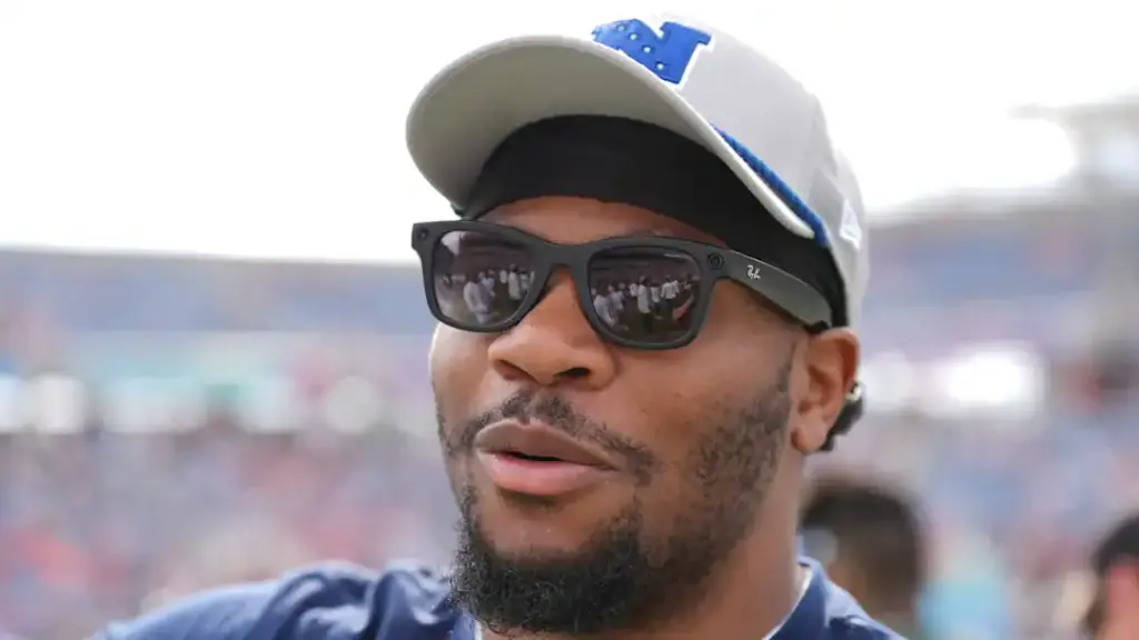 A man wearing sunglasses and a cap at an outdoor event, with a blurred crowd in the background, likely attending an American football game or sports gathering.