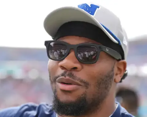 A man wearing sunglasses and a cap at an outdoor event, with a blurred crowd in the background, likely attending an American football game or sports gathering.