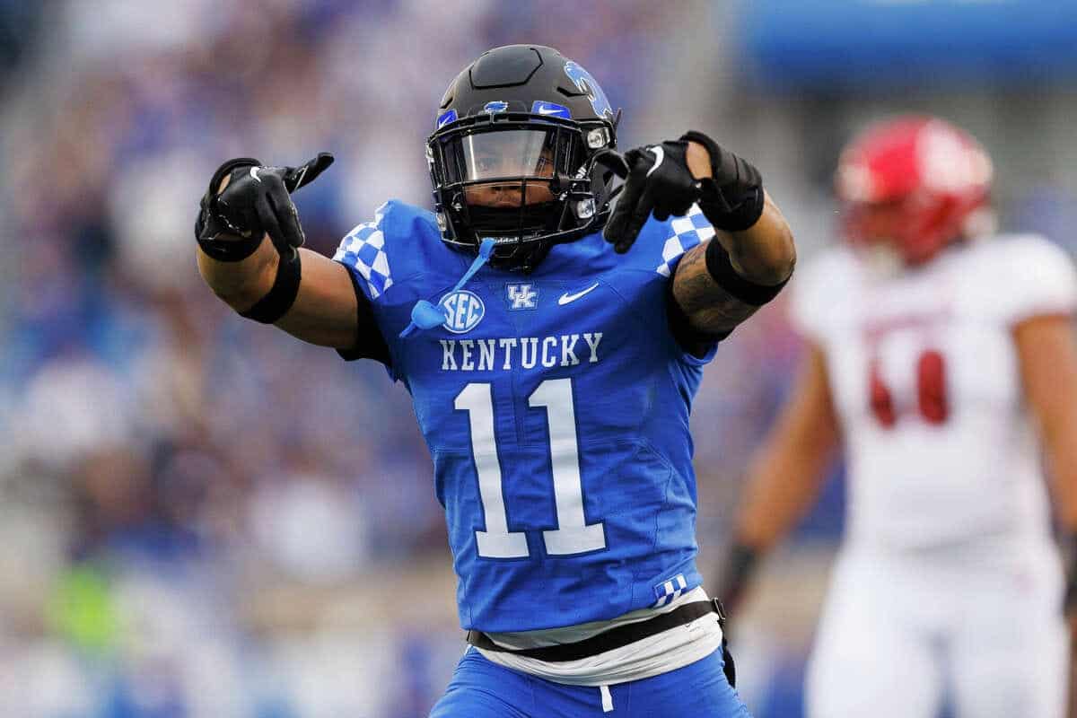 Athletic football player in blue Kentucky Wildcats uniform celebrating on the field, pointing with both hands during a college football game, with a blurred crowd in the background.