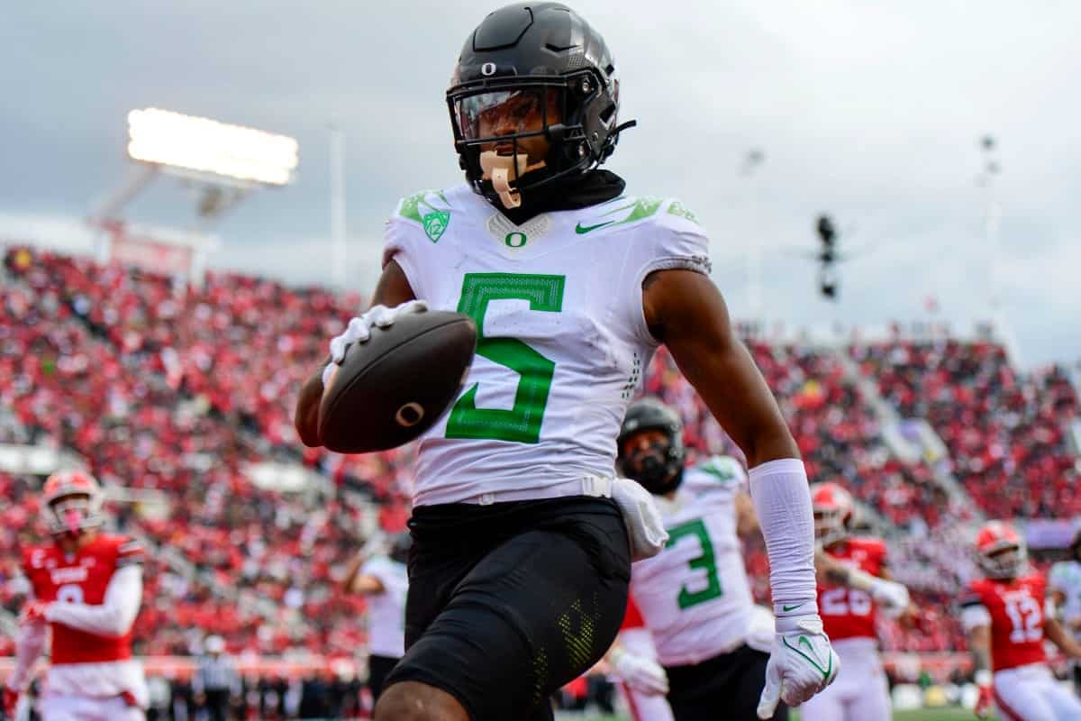 Athlete in football uniform with helmet carrying football during game, fans in stadium background, on-field action scene, college football player, Oregon Ducks, Dallas Cowboys coverage, sports photography.