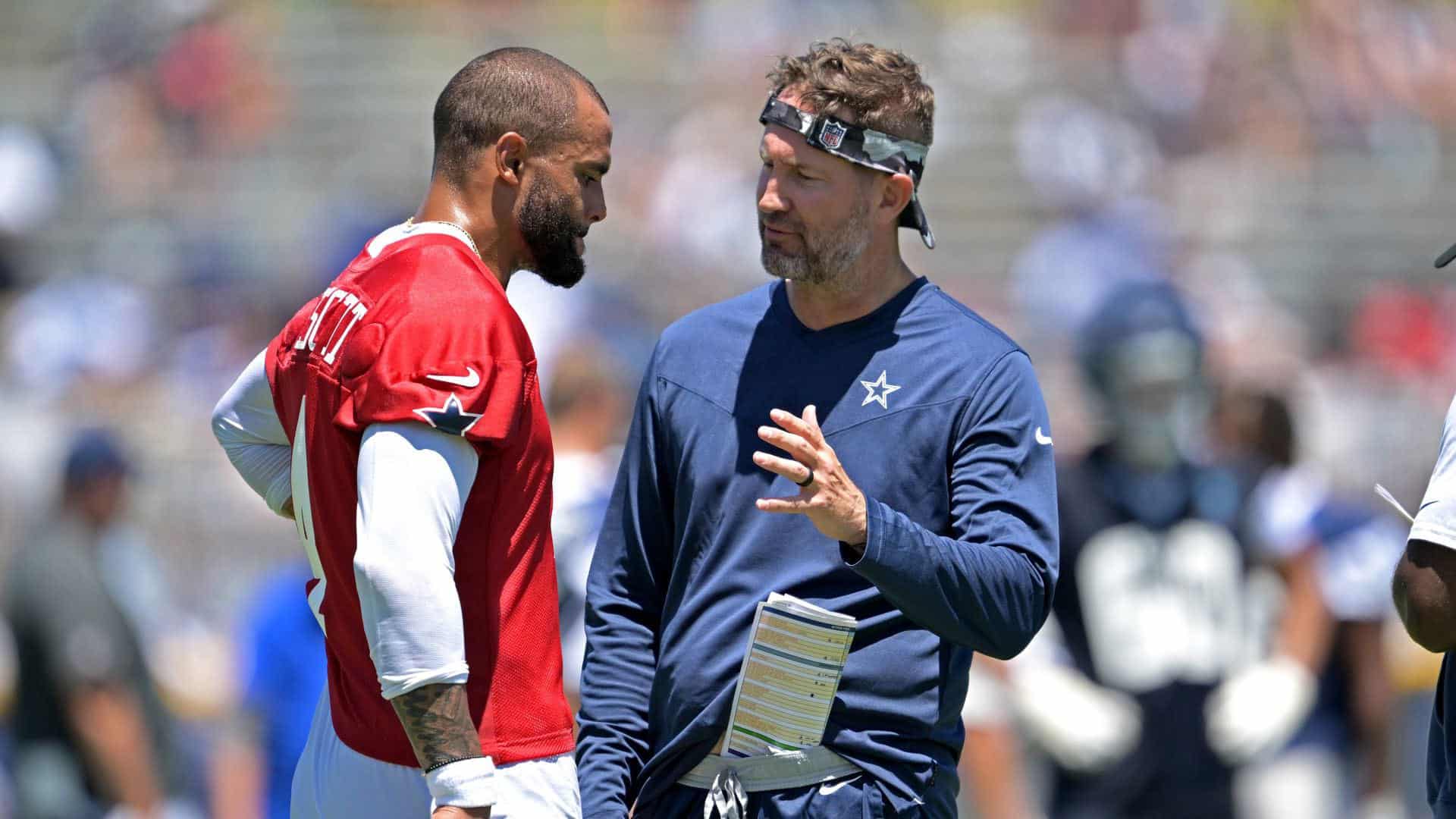 Wide receiver on football field talking with coach, discussing game strategy during NFL practice, Dallas Cowboys player and staff, sunny day, team dynamics, inside the star complex, professional football, sports leadership.