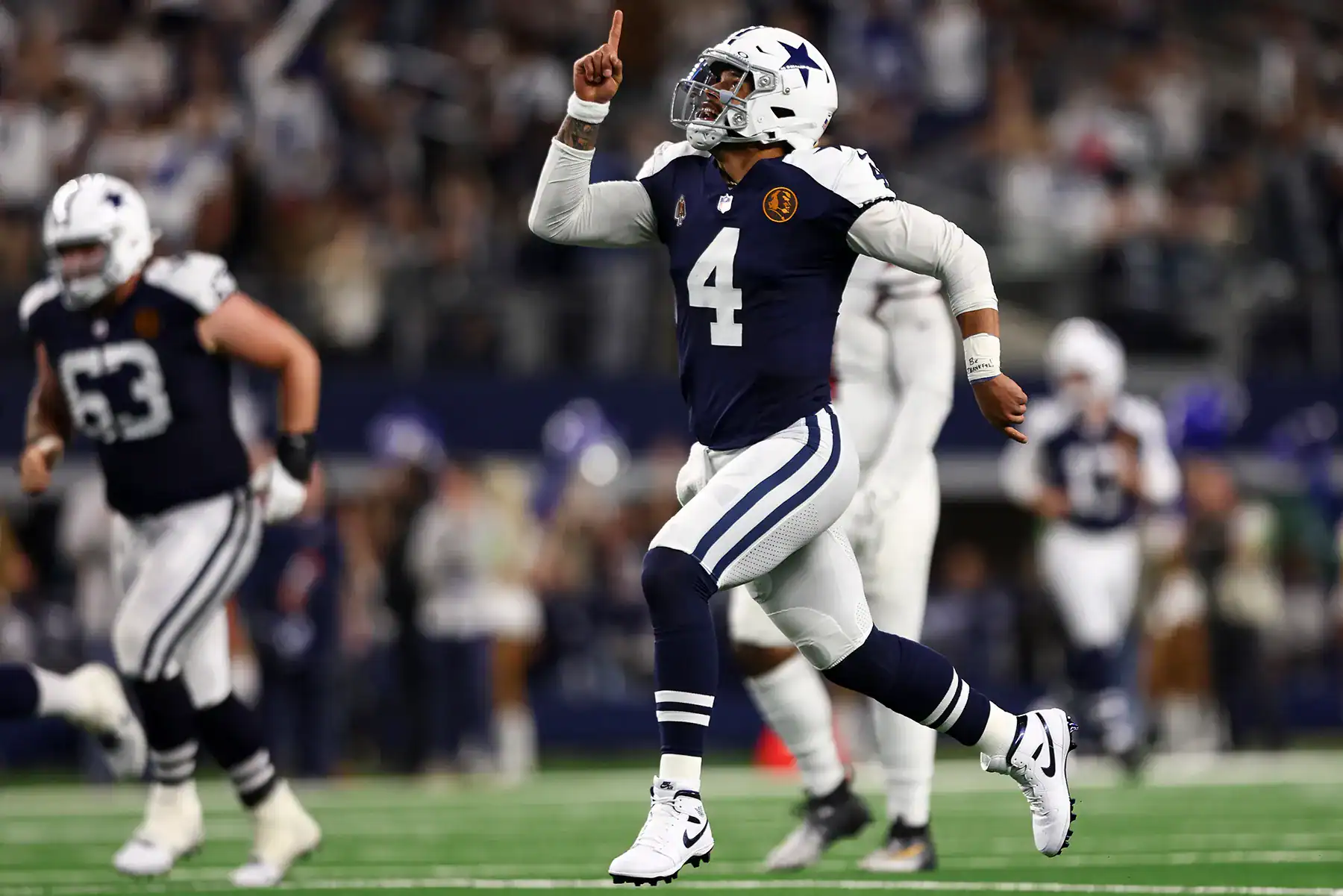 Dallas Cowboys football player celebrating on the field during a game, wearing team uniform and helmet, with fans and teammates in the background.