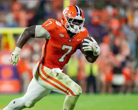 Running football player wearing orange and white uniform holding a football during game.