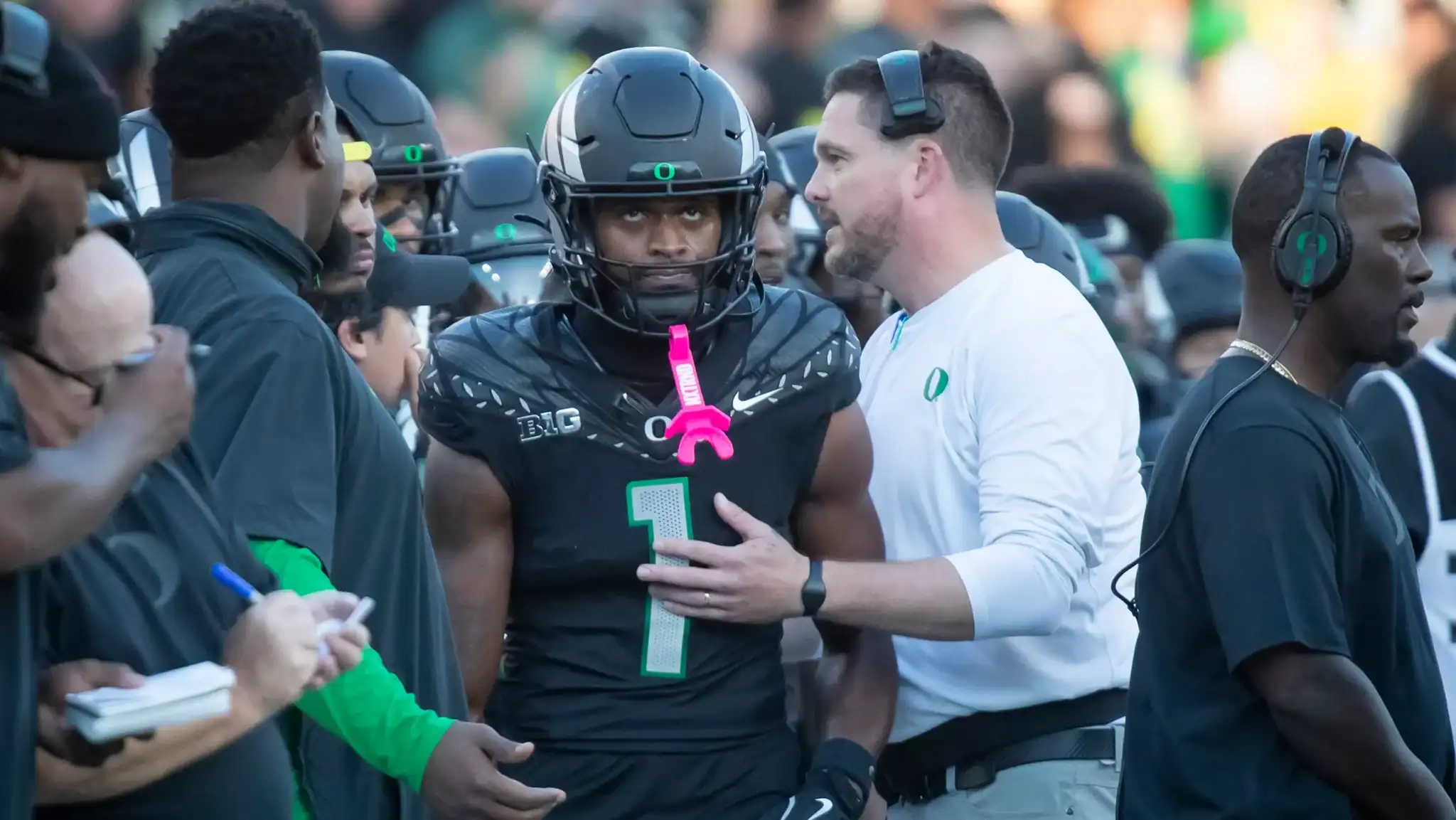 Football player in black uniform with helmet receiving coaching on sideline during game, surrounded by team staff and fans, showcasing Inside The Star's focus on University of Oregon football news and analysis.