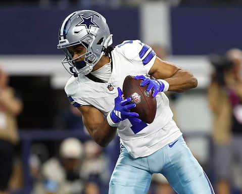 Dallas Cowboys player holding football during game, wearing helmet and white uniform, focused on play, surrounded by blurred crowd, inside the stadium during NFL game.